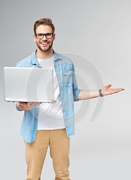 Concentrated young bearded man wearing glasses dressed in jeans shirt holding laptop isolated over grey studio