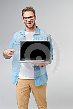 Concentrated young bearded man wearing glasses dressed in jeans shirt holding laptop isolated over grey studio