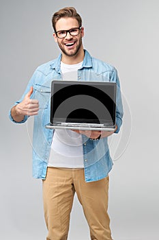 Concentrated young bearded man wearing glasses dressed in jeans shirt holding laptop isolated over grey studio