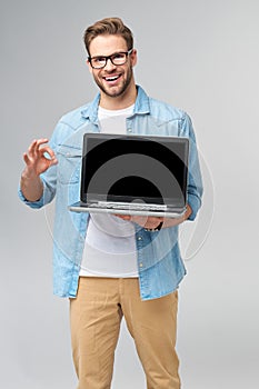 Concentrated young bearded man wearing glasses dressed in jeans shirt holding laptop isolated over grey studio