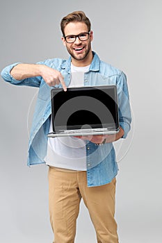 Concentrated young bearded man wearing glasses dressed in jeans shirt holding laptop isolated over grey studio