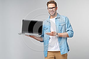 Concentrated young bearded man wearing glasses dressed in jeans shirt holding laptop isolated over grey studio