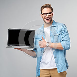 Concentrated young bearded man wearing glasses dressed in jeans shirt holding laptop isolated over grey studio