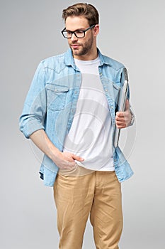 Concentrated young bearded man wearing glasses dressed in jeans shirt holding laptop isolated over grey studio