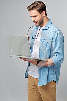 Concentrated young bearded man wearing glasses dressed in jeans shirt using laptop isolated over grey studio background