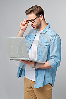Concentrated young bearded man wearing glasses dressed in jeans shirt using laptop  over grey studio background