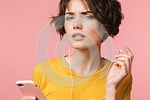 Concentrated young beautiful woman posing isolated over pink wall background listening music with earphones using mobile phone try