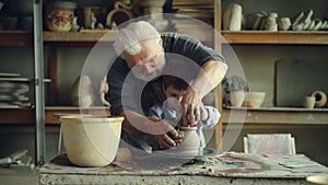 Concentrated young boy is molding clay into ceramic vase on spinning throwing wheel and his experienced grandfather is