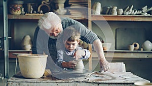 Concentrated young boy is molding clay into ceramic vase on spinning throwing wheel and his experienced grandfather is