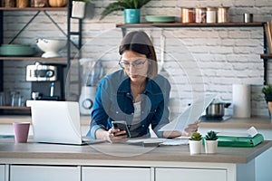 Concentrated young business woman using her mobile phone while working with computer in the kitchen at home