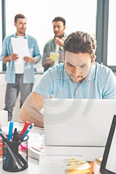 concentrated young businessman using laptop while colleagues standing behind