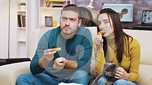 Concentrated young couple sitting on couch eating fried chicken