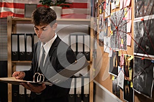 Concentrated young detective in black suit reading dossier in dark office. Investigator working with criminal case