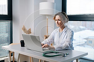 Concentrated young female doctor in white coat typing on laptop computer looking on display screen, sitting at table at