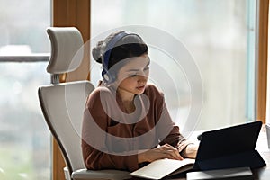 Concentrated young indian woman in headphones listening lecture on touchpad