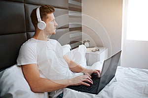 Concentrated young man in bed early morning. He work at home. Guy hold laptop on knees and headphones on ears.