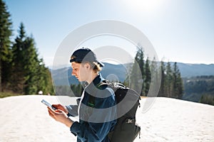 Concentrated young man in a cap and with a backpack on his back and casual clothes standing in the mountains on the snowy slopes