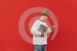 Concentrated young man in casual clothes and headphones walks on the background of a red wall with a laptop in his hands and uses