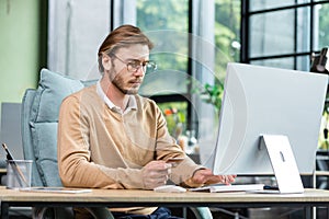 Concentrated young man holds a card in his hands, sits at a desk in the office in front of a computer, makes an online