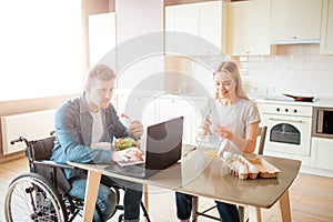 Concentrated young man on wheelchair working with laptop and eating salad. Studying with disability and inclusiveness