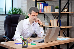 Concentrated young manager man sitting at office desk working on laptop computer and thinking about solving a financial problem