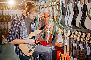 Concentrated young musician stand and play on electric guitar. He looks at it. Guy is in music shop. Young hipster