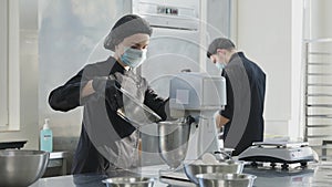 Concentrated young woman in Covid face mask pouring ingredients in automatic mixer with blurred man working on