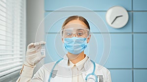 Concentrated young woman doctor in blue disposable face mask and glasses holds vaccine vial in hand