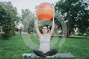 Concentrated young woman sit in lotus pose on yoga mate in park. She hold orange fitness ball up with hands.