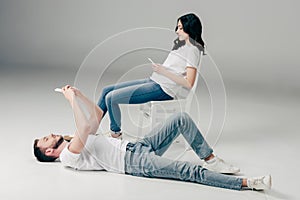 Concentrated young woman sitting on chair and using smartphone near handsome man lying on floor and taking selfie