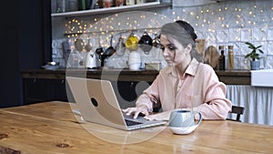 Concentrated young woman sitting in the kitchen at wooden table and typing on her personal silver laptop with the cup of