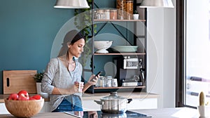 Concentrated young woman using her mobile phone while drinking a cup of coffee in the kitchen at home