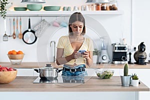 Concentrated young woman using her mobile phone while eating a salad in the kitchen at home