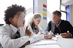 Concentrating Male High School Student Wearing Uniform Working At Table With Teacher Talking To Pupils In Background