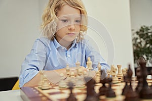 Concentration. Portrait of focused caucasian boy in blue shirt sitting in the classroom and thinking while playing chess