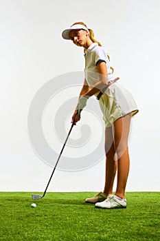 Concentration. Young girl in white sportswear, standing on grass, playing golf isolated over white background
