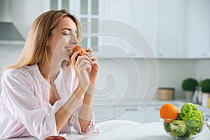 Concept of choice between healthy and junk food. Woman eating croissant at white table in kitchen