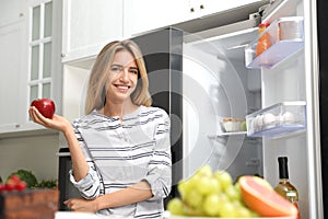 Concept of choice between healthy and junk food. Woman eating fresh apple near refrigerator in kitchen