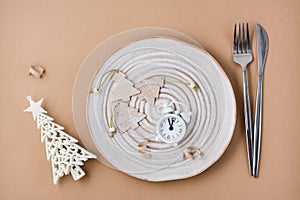 Concept Countdown on Christmas night. Table setting. Alarm clock and Christmas decorations on a plate and cutlery on a beige