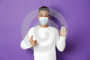 Concept of covid-19, pandemic and social distancing. African-american man in medical mask showing thumbs-up and hand
