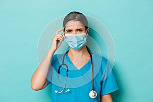 Concept of covid-19 and quarantine concept. Close-up of confused female doctor in medical mask and scrubs, frowning and