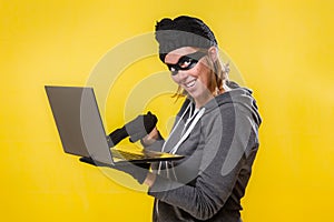 The concept of cybercrime and hacking. Portrait of a woman in a black hat, gloves and mask holding a laptop with a evil smile.