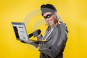 The concept of cybercrime and hacking. Portrait of a woman in a black hat, gloves and mask holding a laptop with a evil smile.