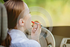 Concept of danger driving. Young woman driver red haired teenage girl painting her lips doing applying make up while driving the