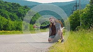 Concept difficulty of traveling alone. Sad desperate hitchhiker man, sits on rural road on sunny summer day. Guy with