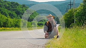 Concept difficulty of traveling alone. Sad desperate hitchhiker man, sits on rural road on sunny summer day. Guy with