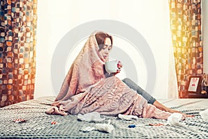 The concept of diseases and flu. A young brunette woman is sitting on the bed, wrapped in a blanket and drinking tea. Next to her