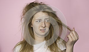 The concept of dry lifeless hair. A woman on a pink background holds her disheveled, tangled hair and looks at it with a