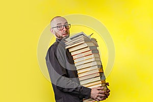 Concept of education. A bald man with a beard and glasses, holding a large stack of books. Yellow background. Copy space