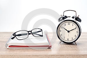 Concept Education or business : The glasses  put on the red book with black alarm clock on wooden table isolated on white